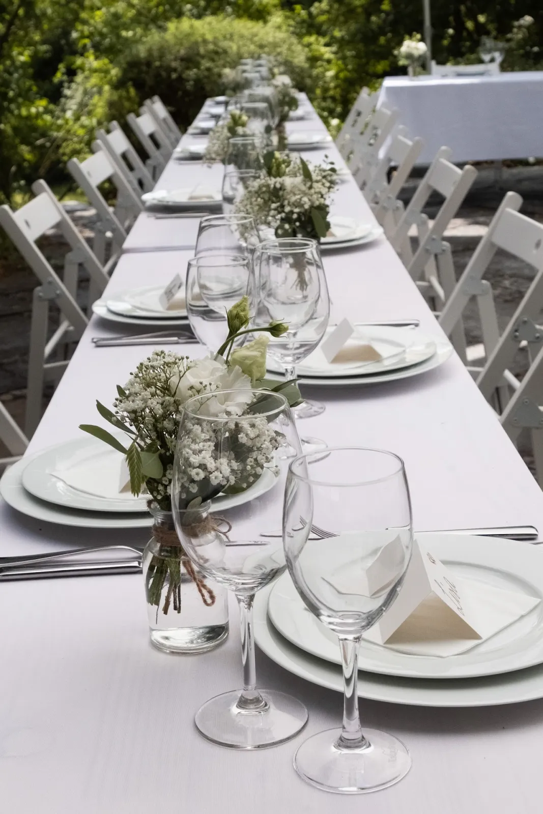 Lange gedeckte Hochzeitstafel mit weißen Holzklappstühlen, Weingläsern und weißen Blumen im Garten